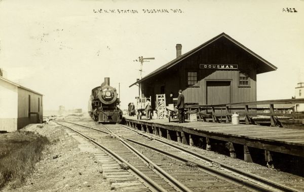 Black and white photographic postcard down two sets of railroad tracks towards the Chicago and Northwestern Railroad station. A number of people are standing on the platform near the locomotive. Caption reads: "C. & N.W. Station, Dousman, Wis."