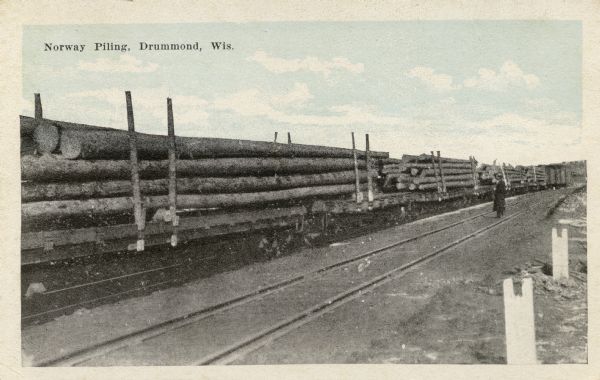 View across railroad tracks towards train flatbeds stacked with large pine logs. There is a man standing further down the tracks looking towards the flatbeds. Caption reads: "Norway Piling, Drummond, Wis."