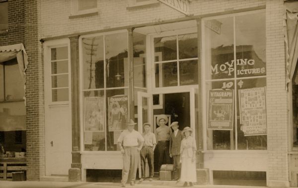 View from across street towards a group of people, four men and a woman,  posing in front of a storefront advertising moving pictures for ten cents. Posters for the movies being shown advertised read: "Roped In," a 1917 release and "Rainbow's End," a 1914 release.