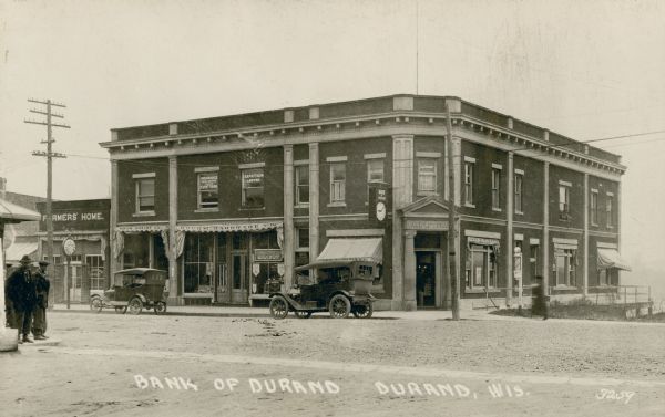 Black and white photographic postcard view from across street towards a two-story brick corner building, labeled as the Bank of Durand. Two Ford Model T's are parked along the curb in front. Pedestrians are standing on the sidewalk on the far left. Caption reads: "Bank of Durand, Durand, Wis."