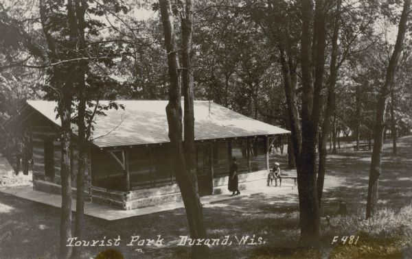 View looking downhill towards a small structure made of logs and with a large screened porch, set in a wooded area. A woman is standing on the walk near the door, and a child is seated on a bench at the right corner of the building. Caption reads: "Tourist Park, Durand, Wis."