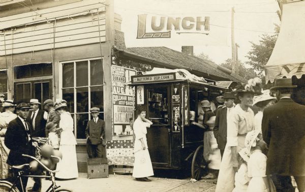 Black and white photographic postcard view of a lunch stand advertising buttered popcorn, ice cream cones and cigars. Groups of men, women and children are gathered on either side of the small stand. A man is walking with his bicycle on the left, and people are lined up at the stand. Visible on the side of a nearby building is a poster advertising a Gollmar Bros Circus Show coming to Durand in August.