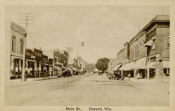 Black and white photographic view down the unpaved Main Street. Includes parked cars, a few pedestrians and commercial buildings. Caption reads: "Main St., Durand, Wis."
