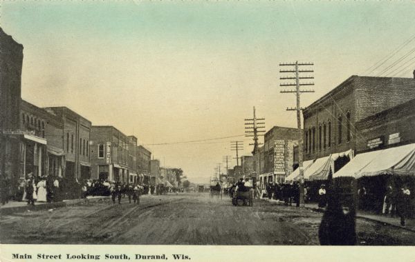 View down center of street looking south. Horse-drawn vehicles are along the street, and telephone poles and many pedestrians line the sidewalks. Caption reads: "Main Street Looking South, Durand, Wis."