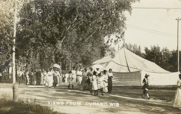 A group of women and girls, some wearing hats or carrying a parasol,  walking along an unpaved street. There is a large canvas tent in the background, with two American flags flying from the top of the tent. There is a group of men gathered behind the women and girls. Caption reads: "Views from Durand, Wis."