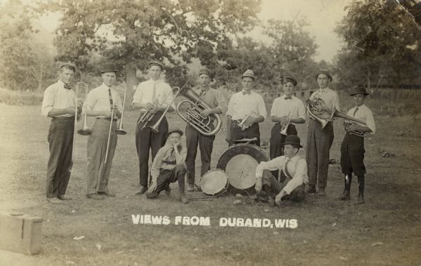 Ten musicians, men and boys, of a small brass band posing with their instruments in an open area outdoors. Includes two drummers. The men are wearing hats, white shirts, neck ties and bow ties.