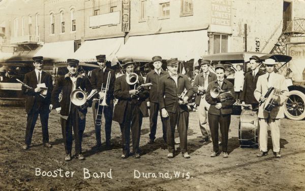 A group of ten musicians posing on an unpaved street with their band instruments. They are wearing hats and band uniforms. Automobiles and commercial buildings are in the background. Caption reads: "Booster Band, Durand, Wis."