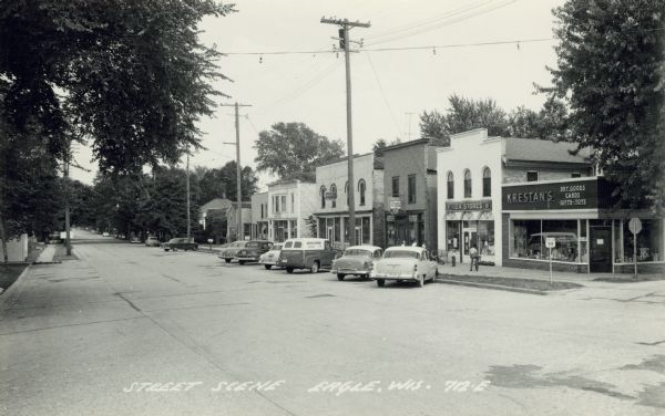 Diagonal view across Hwy 99 going through Eagle, Wisconsin. Probably Jericho Street, but unmarked. Cars are parked at an angle in front of storefronts lining the street on the right. Store signs read: KRESTAN'S Dry Goods, Cards, Gifts, Toys; IGA STORES; Borden's Ice Cream; PATEK PAINTS. Caption reads: "Street Scene, Eagle, Wis."