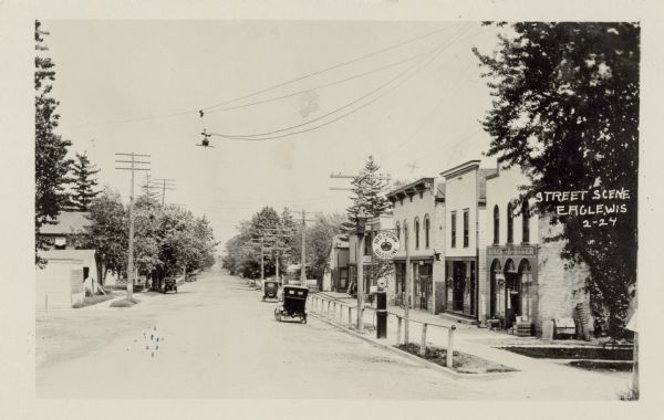 View down center of unpaved street, showing storefronts on the right, with a metal railing between the street and sidewalk. There is a gas pump at the railing with a sign that reads: "Red Crown Gasoline." Three cars are parked further along the street at the curb. Power poles and lines and a streetlight are overhead. Probably Jericho Street. Caption reads: "Street Scene, Eagle, Wis."