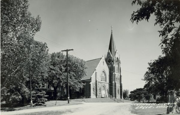 Catholic Church with a spire and stairways in front, on a narrow street. Trees and power poles line the street. Caption reads: "Catholic Church, Eagle, Wis."