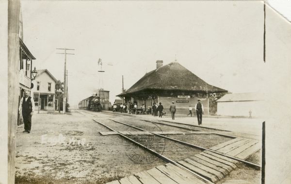 View from left side of double set of railroad tracks towards a locomotive approaching the railroad station from the opposite direction. People are in front of the station, on the right. Other people are walking towards the train. One man on the far left is walking along the side of buildings. There are wooden board walkways crossing the tracks. Caption reads: "Eagle, Wis."
