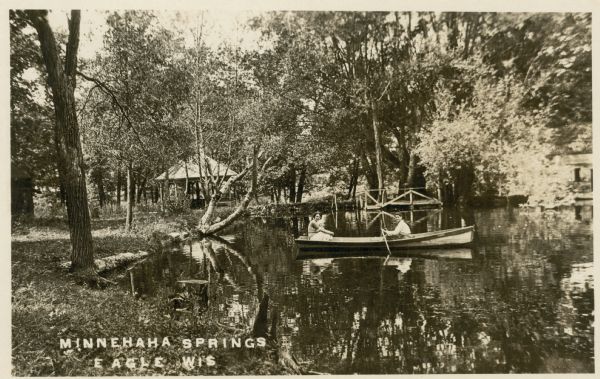 View from shoreline towards a two people in a boat at Minnehaha Springs. In the background is a pavilion among the trees, and a small wooden pier at the shoreline. Caption reads: "Minnehaha Springs, Eagle, Wis."