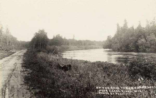 The unpaved road along a river on the way to the Red Oak Resort. Caption reads: "On The Road to Red Oaks Resort, Near Eagle River, Wis."