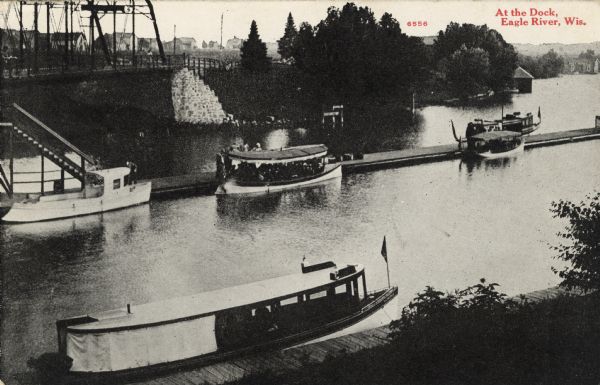 Elevated view of the Eagle River dock, with excursion boats and a tug boat alongside two long piers in the foreground, and a steel frame bridge in the background. Caption reads: "At the Dock, Eagle River, Wis."
