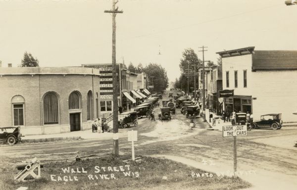 Slightly elevated view of Wall Street (STH 70). In the foreground is an intersection, with a sign that reads: "Look Out for the Cars." There are storefronts on both sides of the street, and automobiles are parked at an angle along the curbs. Pedestrians are standing on the sidewalks. Caption reads: "Wall Street, Eagle River, Wis."