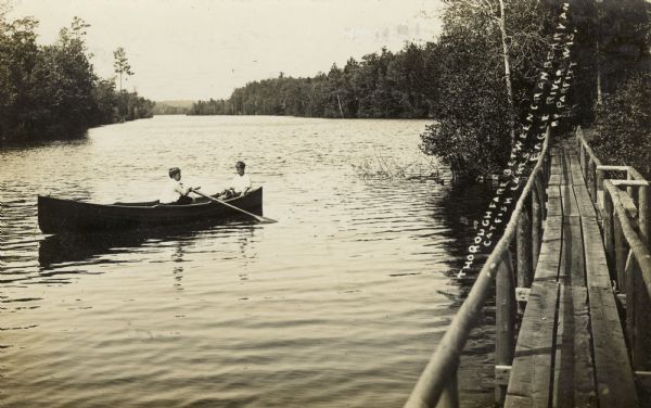 View from pier of two boys in a rowboat in the river, east of Eagle River. Caption reads: "Thoroughfare Between Cranberry & Catfish Lakes, Eagle River, Wis."