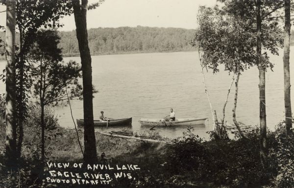 View looking down hill towards two people in rowboats near the shoreline on Anvil Lake, east of Eagle River, on STH 70. Caption reads: "Anvil Lake, Eagle River, Wis."