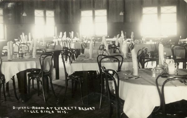 Dining Room of the resort on Catfish Lake, east of Eagle River off of STH 70. The tables are set with white tablecloths and silverware settings. Established in 1896. Caption reads: "Dining Room at Everett Resort, Eagle River, Wis."