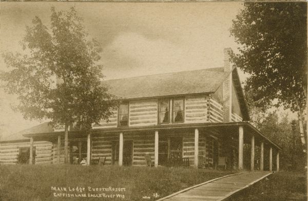 Main Lodge at the resort on Catfish Lake, east of Eagle River off of STH 70. The original lodge was a two-story, log building with a wrap-around porch, and boardwalk leading to the porch. Built in 1896. Caption reads: "Main Lodge, Everetts Resort, Catfish Lake, Eagle River, Wis."