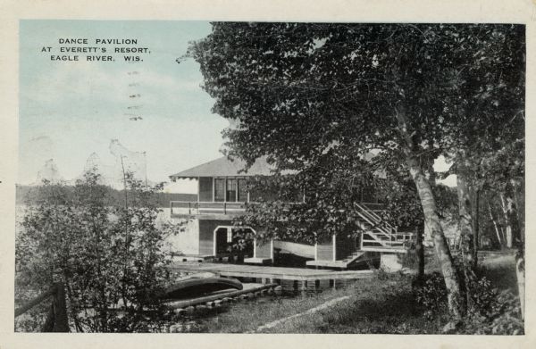 View through trees toward the dance pavilion at Everett's Resort on the shoreline of a lake. Boats are lying on a dock near the shoreline. Caption reads: "Dance Pavilion at Everett's Resort, Eagle River, Wis."