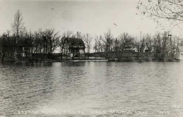 Black and white photographic postcard of three private cottages along the shoreline of Eagle Springs Lake. There is a water tower on the far right. Caption reads: "Dadman, Runkel(?) & Smith, Eagle Springs Lake, Wis."