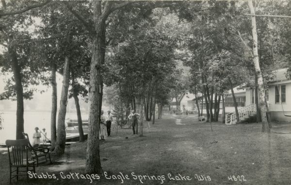 Black and white photographic postcard of Stubbs Cottages on Eagle Springs Lake. People are standing on the lawn along the lakeshore. Caption reads: "Stubbs Cottages, Eagle Springs Lake, Wis."