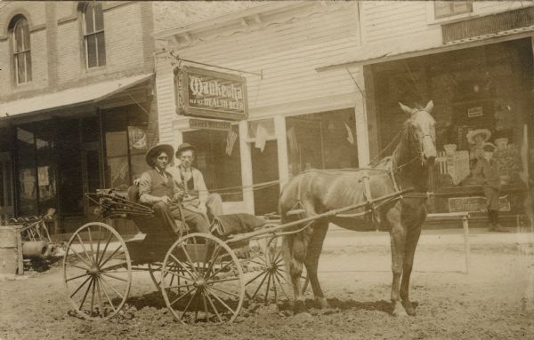 Black and white photographic postcard of two men in a horse-drawn buggy parked in the middle of a unpaved street. Storefronts and pedestrians are behind them along a sidewalk. The sign on the storefront behind them reads: "Waukesha Health Beer."