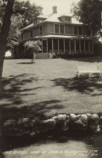 Two-story home with a wrap-around porch. A bench is on the lawn. Caption reads: "Lake Beulah Home of John M. Volkhardts, East Troy, Wis."