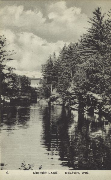Elevated view across Mirror Lake. Trees are along the steep, exposed rock faces along the shoreline. There is a boat near a boathouse on the left, and in the far background is a mill building.