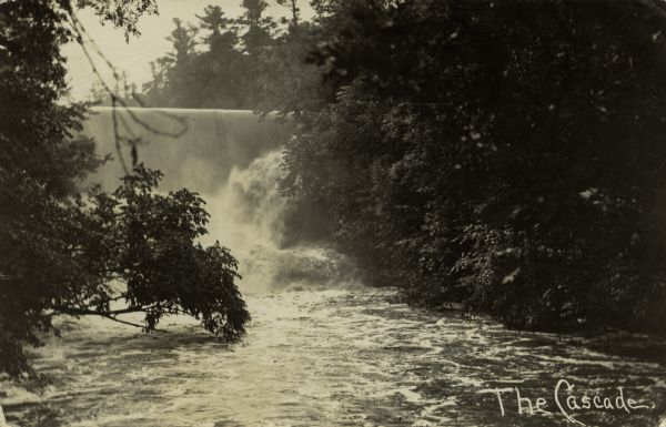 A black and white photographic postcard view looking up a river toward a waterfall. Caption reads: "The Cascades."
