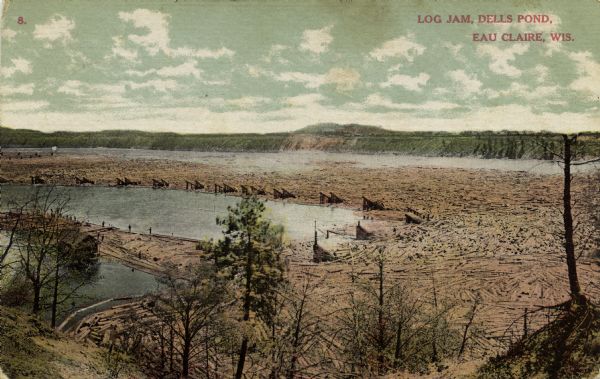 A colored photographic postcard view of a large log jam in the Dells Pond at Eau Claire. There are "booms" and several men standing on logs. Caption reads: "Log Jam in Dells Pond, Eau Claire, Wis."