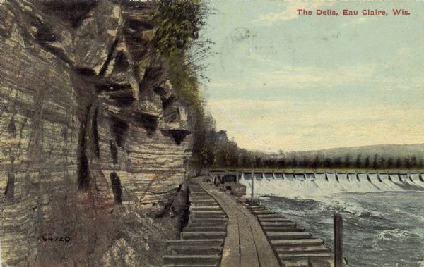 Colored photographic postcard view of the dells below the dam on the Chippewa river. There is a boardwalk at the base of the cliff leading to the dam. Caption reads: "The Dells, Eau Claire, Wis."