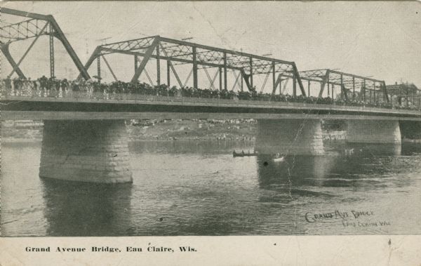 View across river towards a crowd lining the steel truss Grand Avenue Bridge over the Chippewa River. A few rowboats and canoes are on the river. Caption reads: "Grand Avenue Bridge, Eau Claire, Wis."