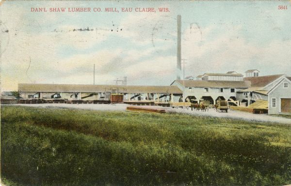 Colorized postcard view across field toward the lumber mill. Caption reads: "Dan'l Shaw Lumber Company Mill, Eau Claire, Wis."