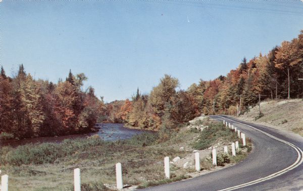 Photographic postcard view of winding road along a river, with trees in fall color.