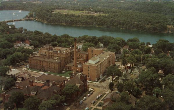 Aerial view of the hospital along the banks of a river, with a bridge on the far left. Beyond the river is a baseball park. On the back of the card is written: "Luther Hospital, 310 Chestnut Street... This non-sectarian, not-for-profit, accredited general hospital serves Eau Claire and the surrounding area, and maintains a School of Nursing.