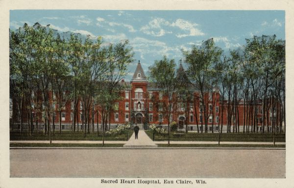 Colorized view of the front of Sacred Heart Hospital. Flowers flank the entrance walk where a man is standing, and a fence is along the sidewalk in front. Caption reads: "Sacred Heart Hospital, Eau Claire, Wis."