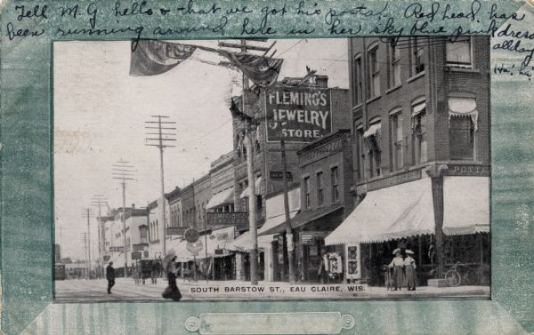 Black and white photographic postcard with a green border. The view is towards the block of South Barstow Street, with a large sign for Fleming's Jewelry Store. Caption reads: "South Barstow Street, Eau Claire, Wis."