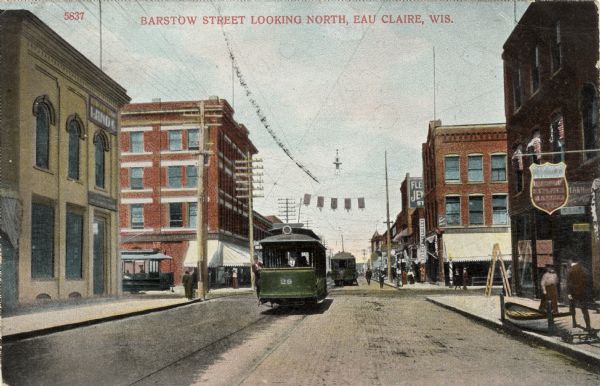 View of the streetcars on Barstow Street, with commercial buildings and pedestrians. There is a boat on the sidewalk on the right. Caption reads: "Barstow Street Looking North, Eau Claire, Wis."