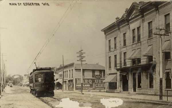 View down Main Street towards a streetcar coming up the street. On the opposite side of the street are commercial buildings. The Monarch Building is in the center background. Caption reads: "Main St., Edgerton, Wis."