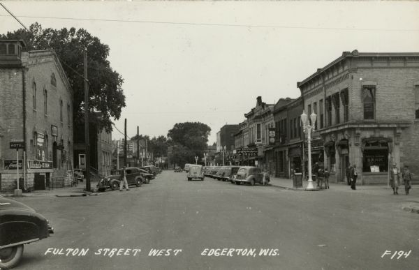 View down center of Fulton Street which is lined with street lamps, parked cars, and commercial buildings. Pedestrians are strolling along the street and sidewalks. Caption reads: "Fulton Street West, Edgerton, Wis."