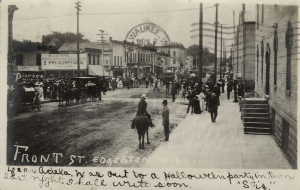 Slightly elevated view of a parade on Front Street. Crowds of people are watching from the sidewalks. Caption reads: "Front St., Edgerton, Wis."