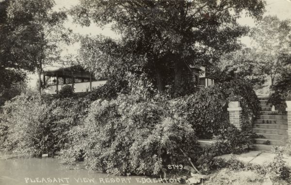 View from water towards a building with a large porch obscured by trees. A flight of steps bordered by two brick columns leads from the lawn to the shoreline. Caption reads: "Pleasant View Resort, Edgerton, Wis."