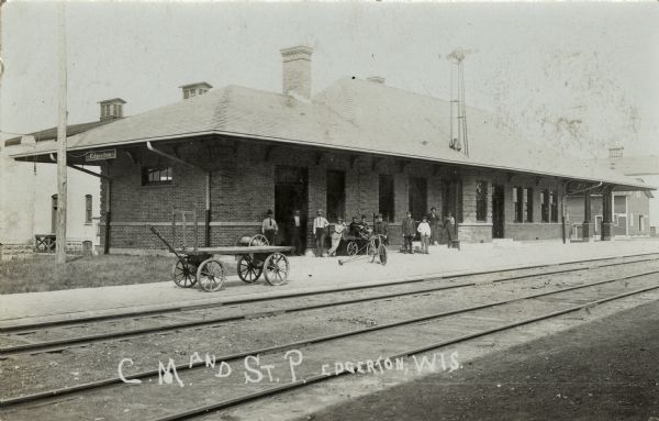 View across railroad tracks towards the station. People and push carts are along the platform. One man has a bicycle. Caption reads: "C.M. and St. P. Edgerton, Wis."