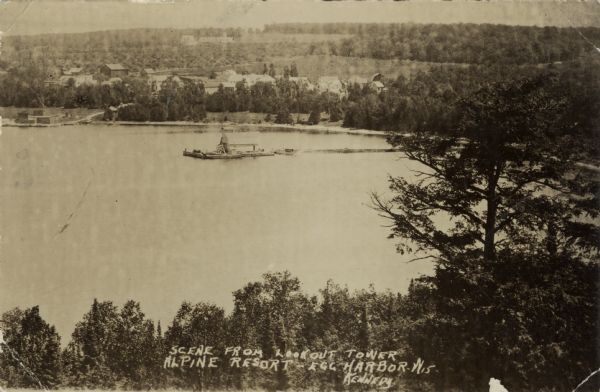 Elevated view of Egg Harbor from the Lookout Tower at Alpine Resort. In the water is a long pier leading to a barn. Dwellings are along the shoreline in the background. Caption reads: "Scene From Lookout Tower, Alpine Resort - Egg Harbor, Wis."