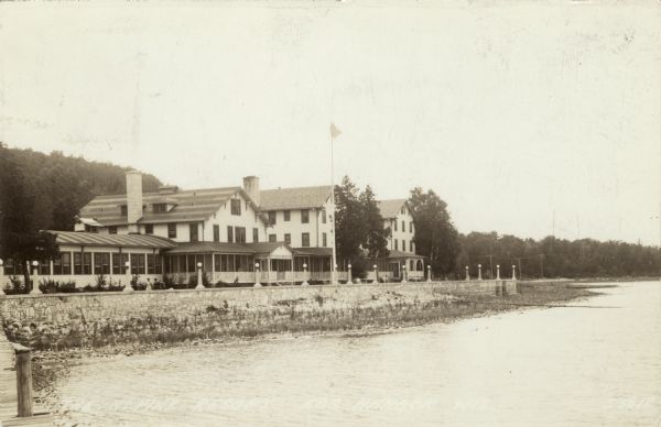 View from pier across water towards the Pine Resort, which is a three-story building with screened-in porches on the shore of Egg Harbor. Street lamps line the top of a stone wall along the shoreline. Caption reads: "The Pine Resort, Egg Harbor, Wis."