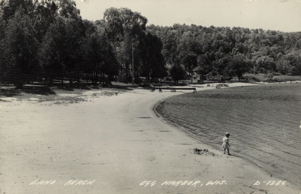 View across beach towards a small boy standing at the water's edge. Rowboats are resting on the shoreline in the background. Caption reads: "Sand Beach, Egg Harbor, Wis."