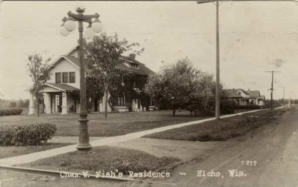 Photographic postcard view of a wood-frame home with young trees in the large yard. A lamppost is on the corner. Other homes are further down the block. Caption reads: "Chas. W. Fish's Residence, Elcho, Wis."