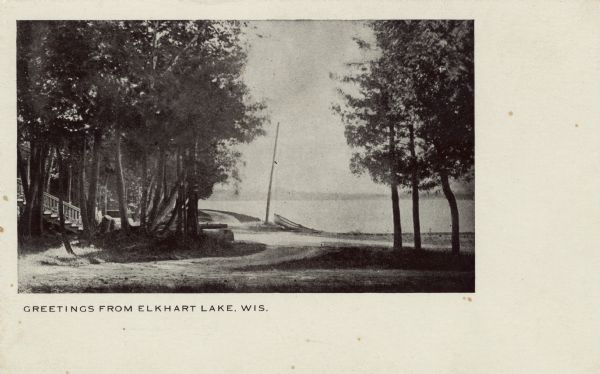 View of the lake from a dirt road. Dwellings are on the left. A small boat is pulled up on the shoreline.