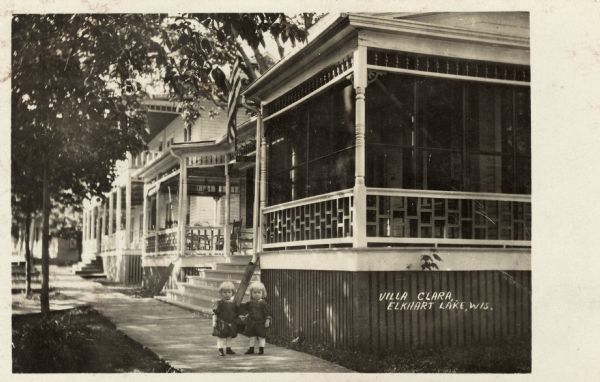 A home with a screened-in porch at the end of a row of houses. Young twins are standing on the sidewalk in front. Caption reads: "Villa Clara, Elkhart Lake, Wis."
